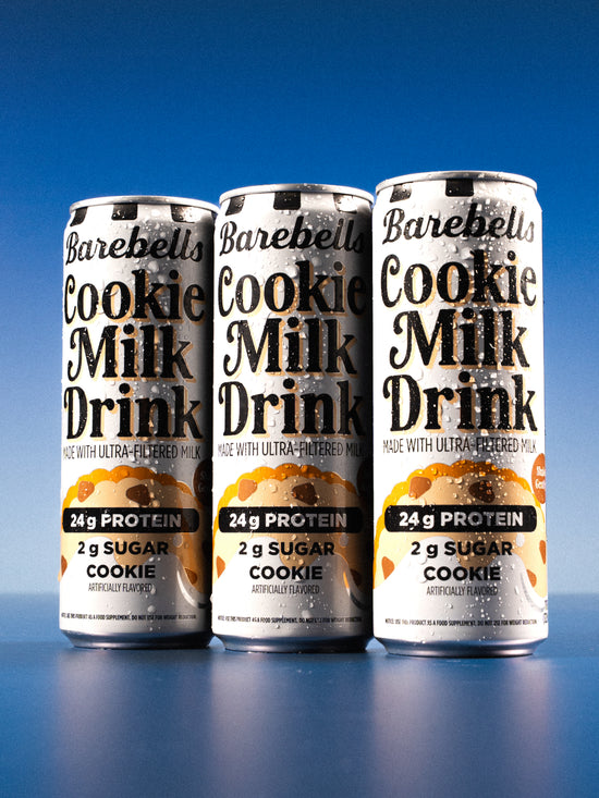 Three cans of Barebells Cookie Milk Drink with condensation, showing 24g protein and 2g sugar per serving against a blue background