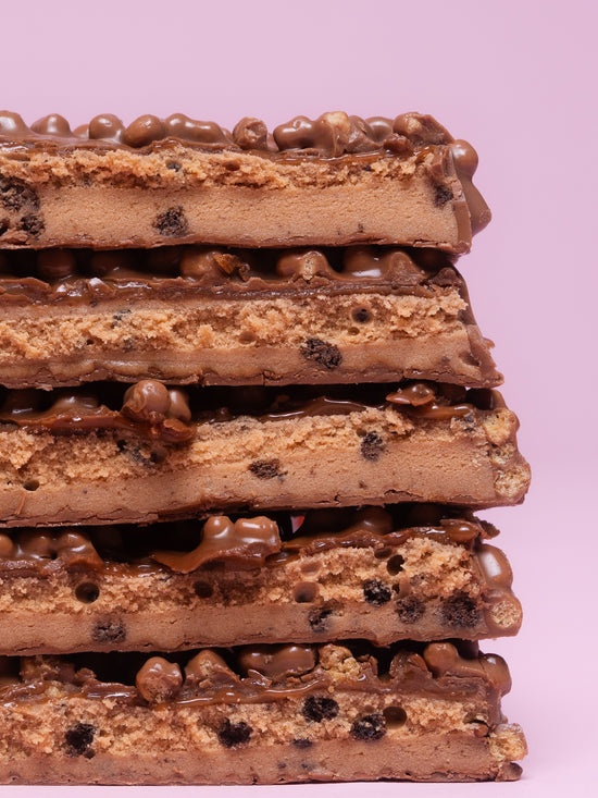 Stack of six Barebells Cookies and Caramel protein bars showing cross-sections with cookie pieces, caramel filling and chocolate coating on pink background
