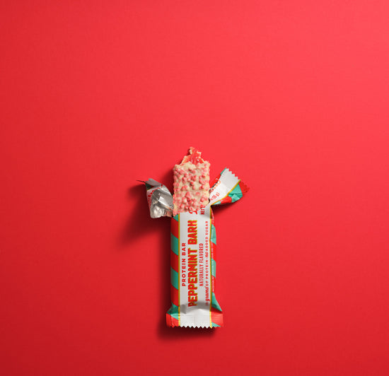 Barebells Peppermint Bark protein bar with white, red and green wrapper, partially unwrapped to show pink and white speckled bar interior against red background