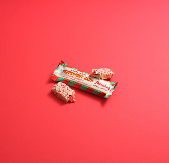 Barebells Peppermint Bark protein bar with white and red striped packaging, displayed with two unwrapped bars showing pink and white speckled coating against a coral background