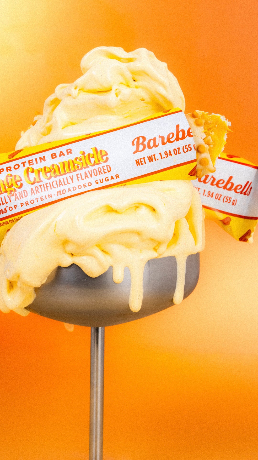 Barebells Orange Creamsicle protein bar on a stick with vanilla and orange swirl coating against an orange gradient background