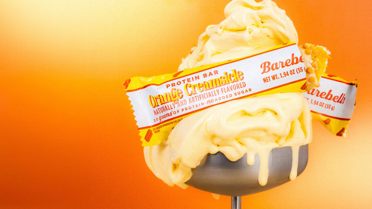 Barebells Orange Creamsicle protein bar on top of a swirled ice cream cone against an orange gradient background
