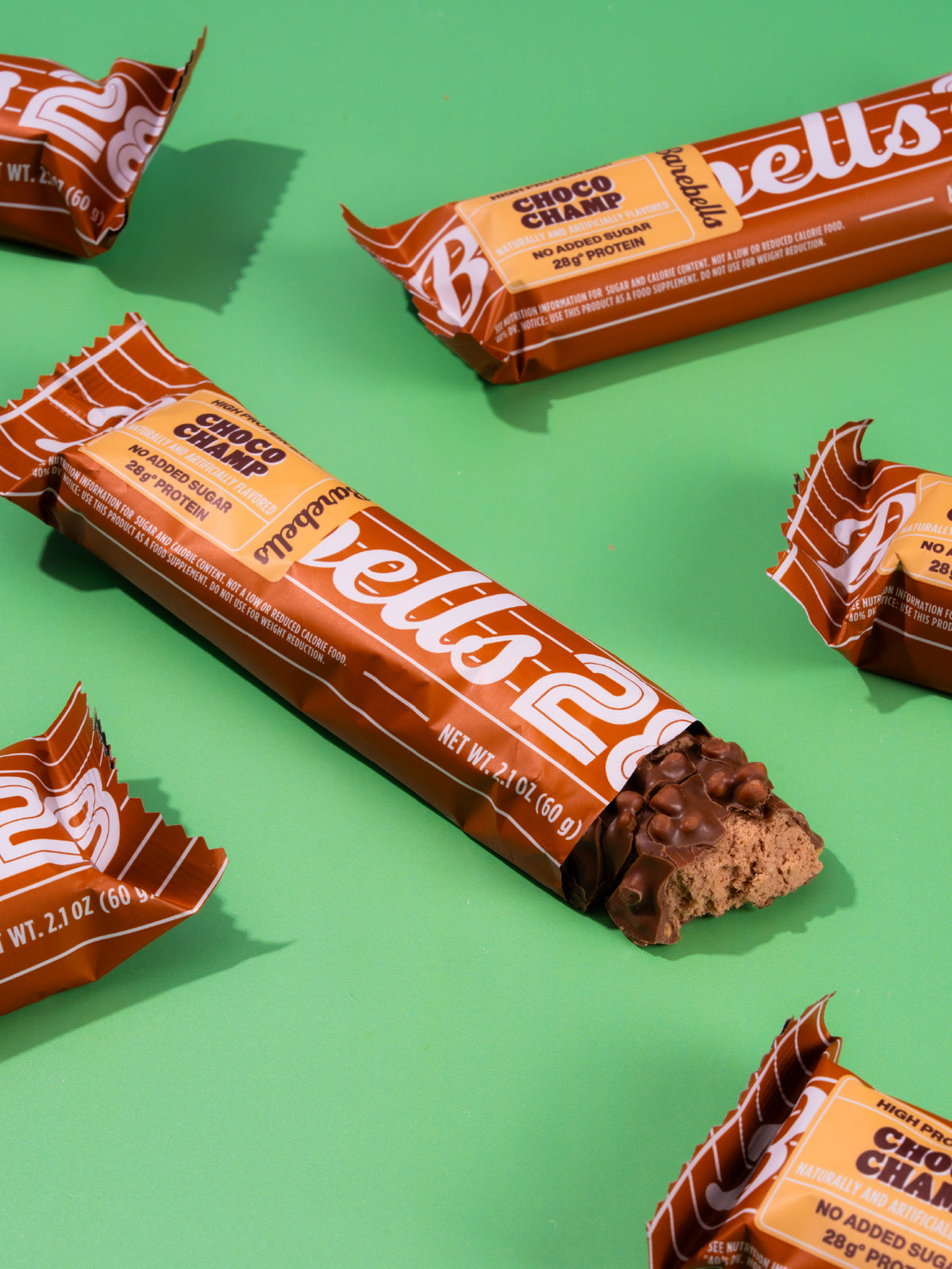 Barebells Choco Champ protein bars with orange and brown packaging arranged on green background, one bar unwrapped showing chocolate filling